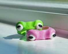 two toy cars with googly eyes sitting on the window sill, one pink and one green