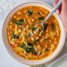 a person is holding a spoon over a bowl of soup with noodles and spinach