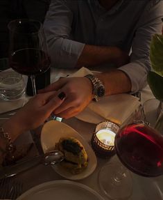 two people sitting at a table with food and wine in front of them, one holding the other's hand