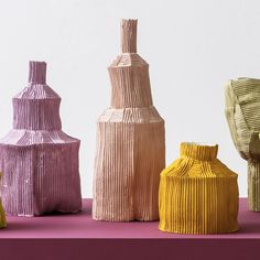 five different colored vases sitting on top of a pink tableclothed surface with white walls in the background