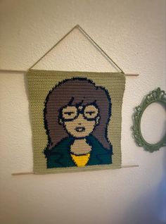 a crocheted picture hanging on the wall next to a frame with a picture of a woman wearing glasses