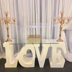 a glass table topped with two candles and a love sign in front of white drapes