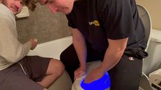 two men are sitting on the toilet and one is touching the lid with his hand