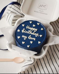 a blue birthday cake with white hearts on it sitting in a box next to a spoon