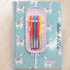 three pens in a pink case on top of a notebook with unicorns and hearts