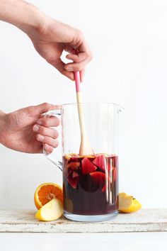 two hands are holding a wooden spoon over a pitcher filled with liquid and sliced oranges