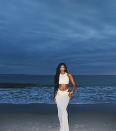 a woman standing on top of a sandy beach next to the ocean at night with her hands in her pockets