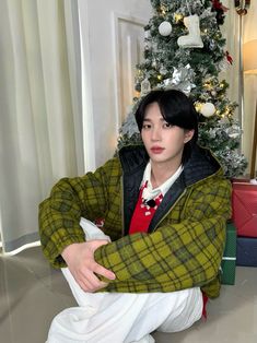 a young man sitting in front of a christmas tree wearing white pants and a green jacket