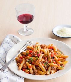 a white plate topped with pasta and meat next to a glass of red wine on top of a wooden table