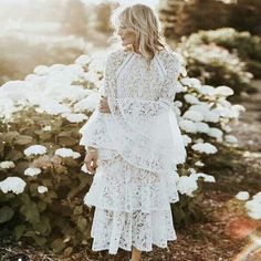 a woman standing in front of flowers wearing a white dress with long sleeves and tiered skirt