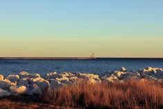 a large body of water sitting next to some rocks and dry grass with a lighthouse in the distance