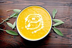 a bowl of carrot soup on a wooden table with green leaves around it and the letter g painted in white