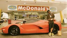 a man standing next to an orange sports car in front of a mcdonald's