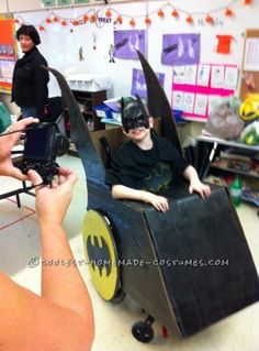 a young boy in a batman costume sitting on top of a toy car while someone takes a photo