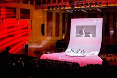 a large screen in the middle of an auditorium with people sitting on chairs around it