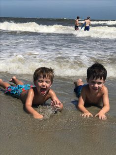 two young boys laying on their stomachs in the water at the beach while one boy watches