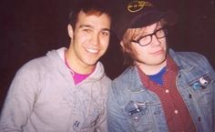 two young men standing next to each other in front of a dark background, one wearing a hat and the other smiling