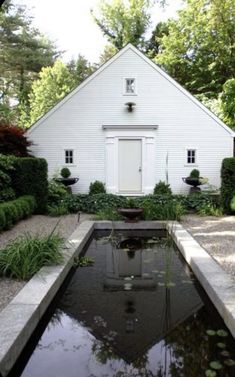 a small white house with a pond in front of it and lots of greenery