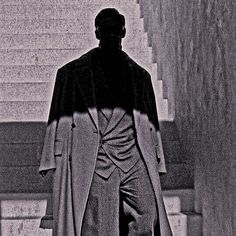 a black and white photo of a man in a long coat with his back turned to the camera