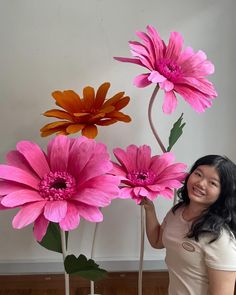 a woman standing next to two large pink and orange flowers in a vase on top of a wooden floor