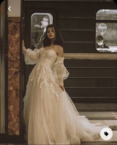 a woman standing in front of a train wearing a wedding dress with long sleeves and an off the shoulder design