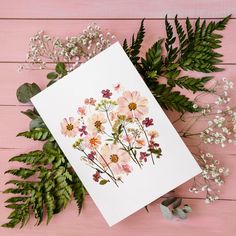 a card with pink flowers and greenery on a pink wooden table next to it