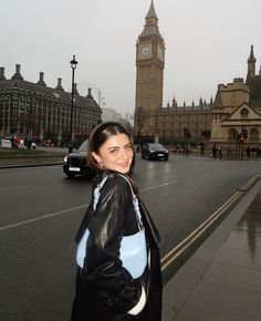 a woman standing on the side of a road in front of a clock tower and buildings