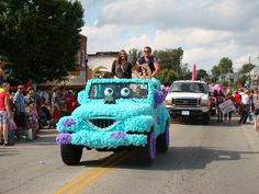 two people riding on the back of a blue car in a parade with other people watching