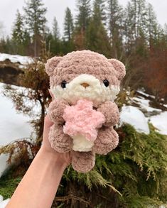 a hand holding a small stuffed animal in front of snow covered ground and evergreen trees