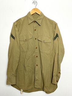 This shirt has marks on both sides and on the sleeve. It also shows distressing and wear on the cuff. (See photos for flaws) Measurements  Pit to pit (width): 21 1/2 inches Top to bottom (length): 28 inches Retro Khaki Button-up Shirt, Military Style Long Sleeve Cotton Shirt, Military Style Cotton Long Sleeve Shirt, Retro Long Sleeve Khaki Top, Vintage Khaki Tops With Snap Buttons, Cotton Uniform Shirt With Long Sleeves, Retro Khaki Long Sleeve Top, Cotton Long Sleeve Uniform Shirt, Vintage Khaki Button-up Shirt