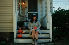 a woman sitting on the front steps of a house with pumpkins and gourds