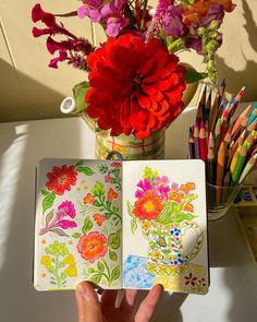 a hand holding an open notebook with colorful flowers in it and colored pencils on the table