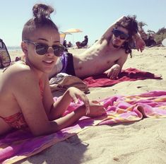 two people laying on the beach with towels in front of them and one person wearing sunglasses