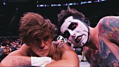 two wrestlers with their faces painted white and one is wearing a clown mask while the other has his arm wrapped around him