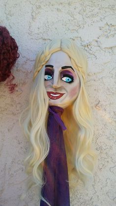 a doll with blonde hair and blue eyes wearing a purple neck tie next to a white wall