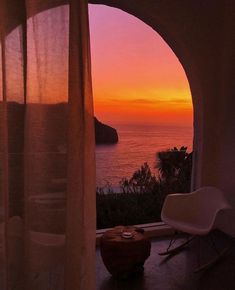 the sun is setting over the ocean from an arched window with sheer drapes on it