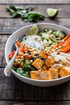 a white bowl filled with vegetables and tofu on top of a wooden table next to lime wedges