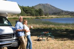 a man and woman standing next to a camper van with a lake in the background