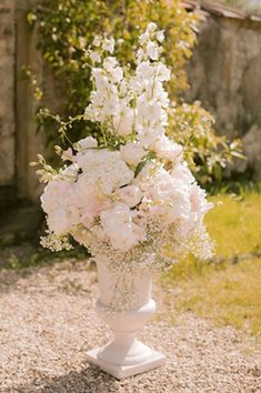 a vase filled with white flowers sitting on top of a gravel road next to a stone building