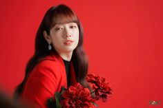 a woman with long hair holding flowers in front of a red wall and looking at the camera