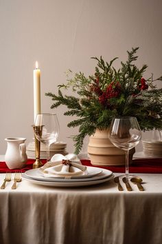 a table set for christmas dinner with white plates and silverware, candlesticks and greenery