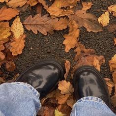 a person's feet in jeans and black shoes on the ground surrounded by leaves