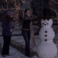 two women standing next to a snowman in the middle of a snowy street at night