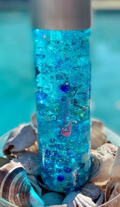 there is a glass vase with blue liquid in it on top of the sand and shells