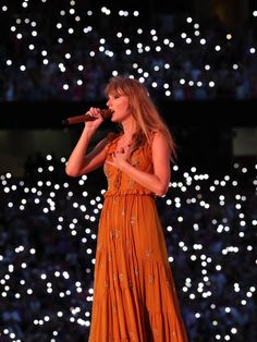 a woman in an orange dress holding a microphone and singing into a microphone with lights behind her