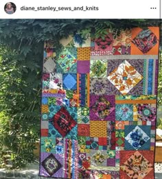 a colorful quilt hanging from the side of a tree in front of some bushes and trees