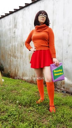 a woman in an orange sweater and red skirt holding a shopping bag while standing on grass