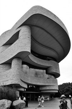 black and white photograph of people walking in front of a large building with curved roof