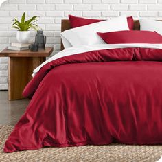 a bed with red sheets and pillows in front of a white brick wall next to a table