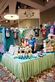 two people standing behind a table with items for sale on it in front of a display